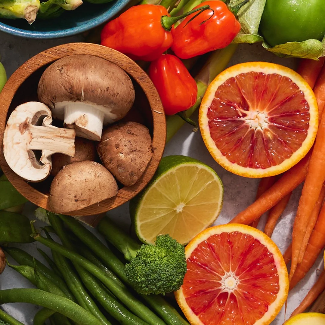 Assorted fresh fruits and vegetables, including blood oranges, a lemon, capers, green beans, carrots, shiitake mushrooms, cherry peppers, broccoli, and a green apple.