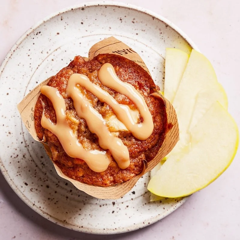 A muffin topped with drizzled icing on a speckled ceramic plate, accompanied by two slices of yellow apple.