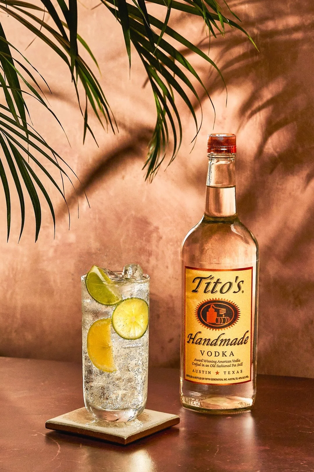 A bottle of Tito's Handmade Vodka and a glass of vodka with lemon slices and lime wedge, set on a wooden surface with a pink background and green palm leaves overhead.