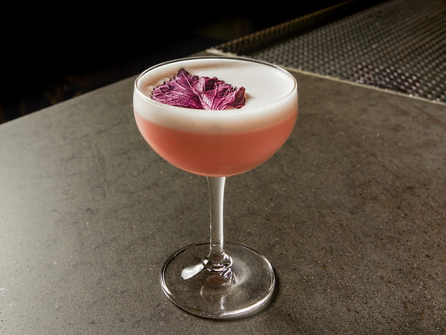 A pink cocktail in a coupe glass topped with a white foam and garnished with a purple autumn leaf on top, placed on a grey surface.