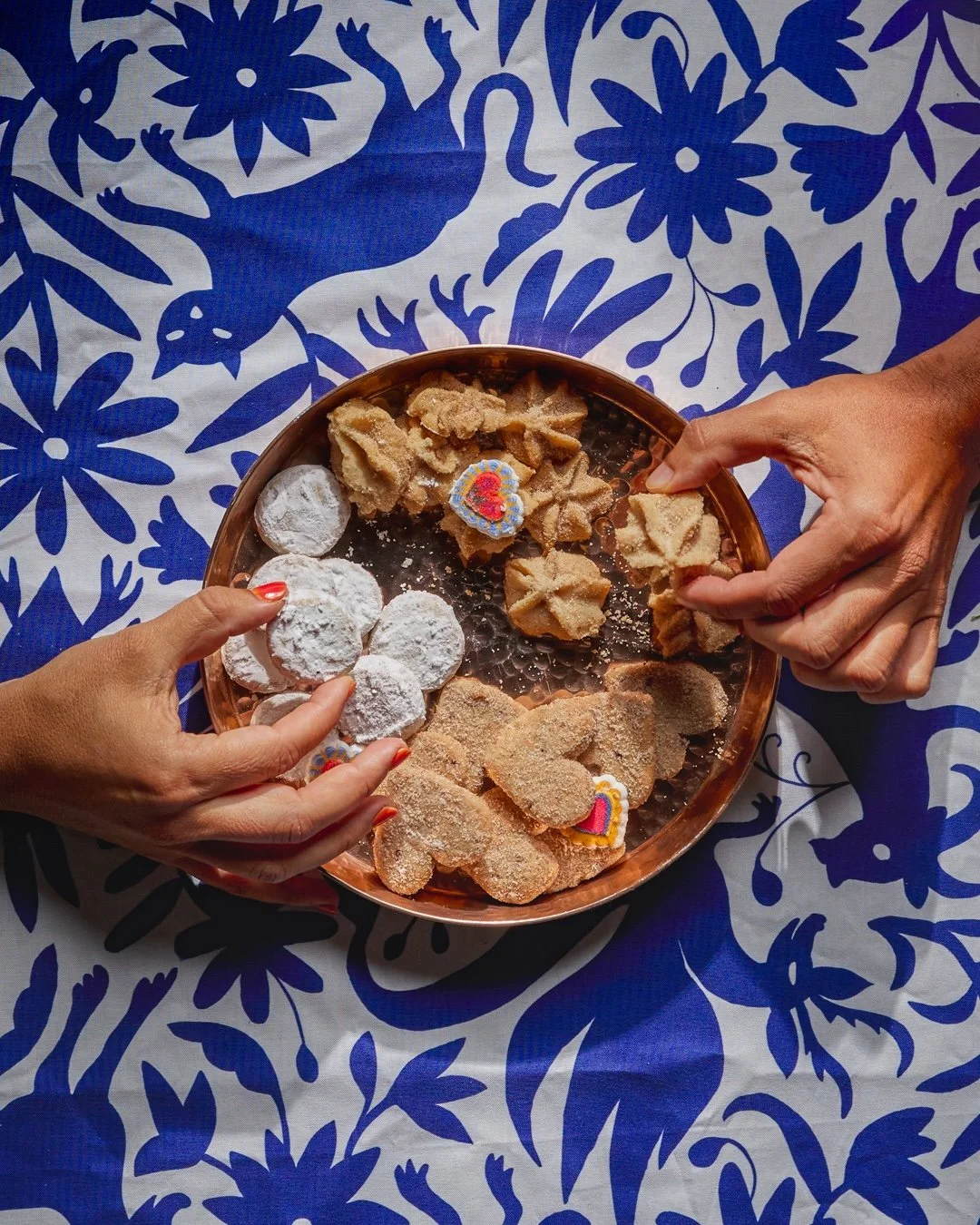 Hands holding a plate of assorted traditional cookies on a blue and white floral tablecloth.