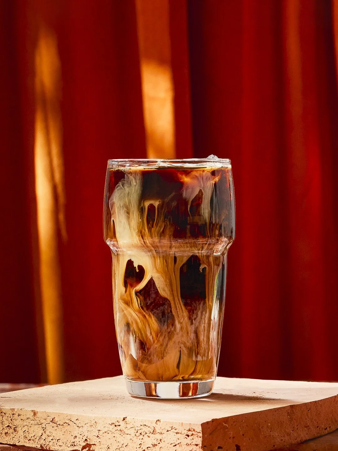 A tall glass of iced coffee with cream swirling in the dark coffee, placed on a wooden surface with a reddish curtain in the background.