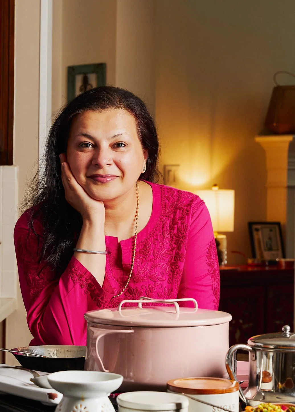 A woman with dark brown hair, wearing a pink embroidered top, sitting at a table with kitchenware, smiling and resting her face on her hand in a warmly lit room.