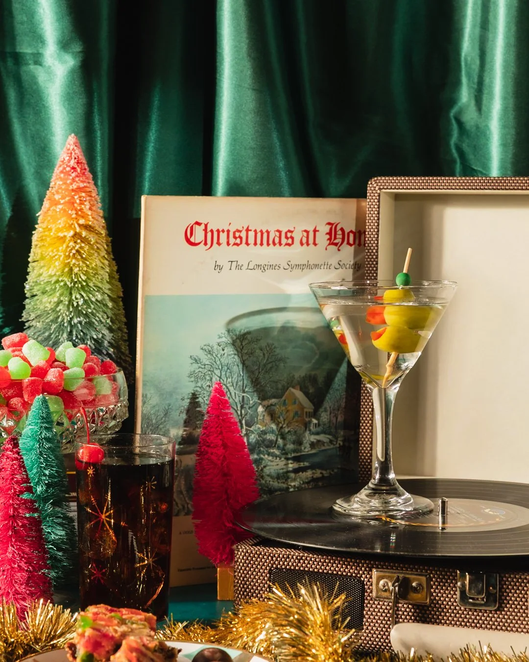 Christmas-themed decorations including a vintage record player with a Martini glass on top, colorful miniature Christmas trees, a bowl of holiday candies, and a book titled "Christmas at Home" set against green curtains.