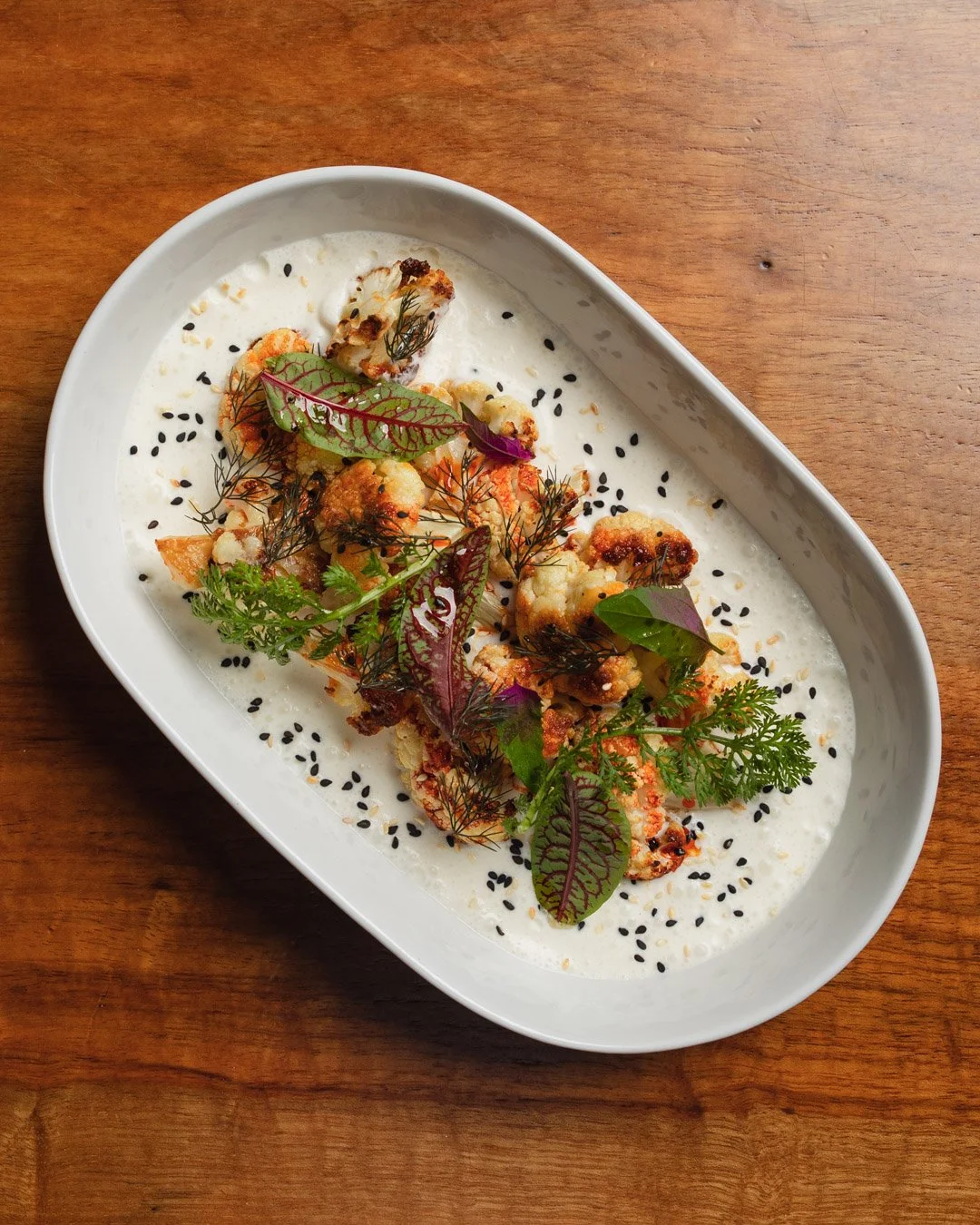 A white oval dish containing roasted cauliflower florets garnished with fresh herbs and microgreens, sprinkled with black sesame seeds, on a wooden surface.