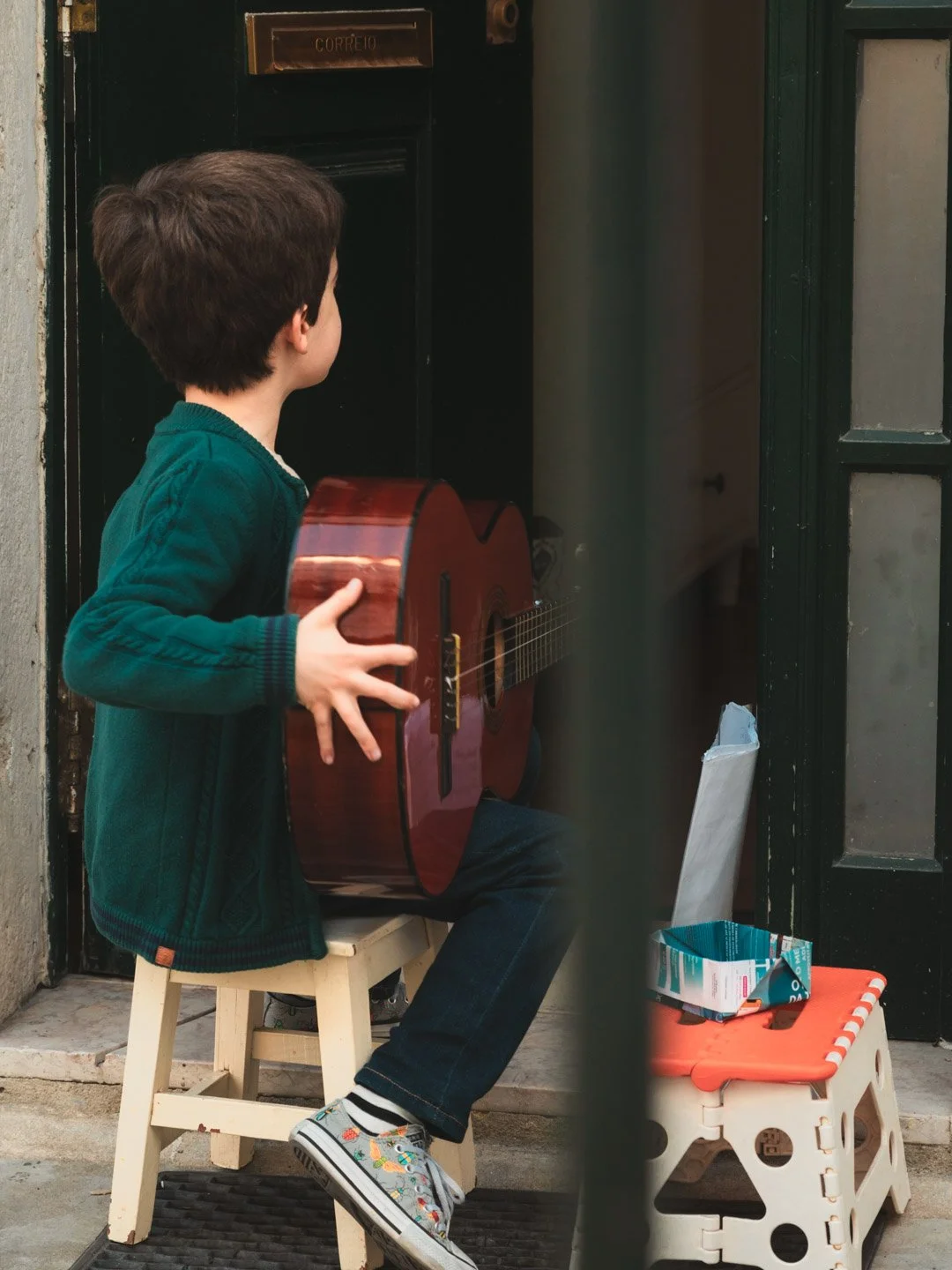 A young boy sitting on a small wooden stool, holding a guitar, looking towards a door with a mailbox label, outside near a building entrance.