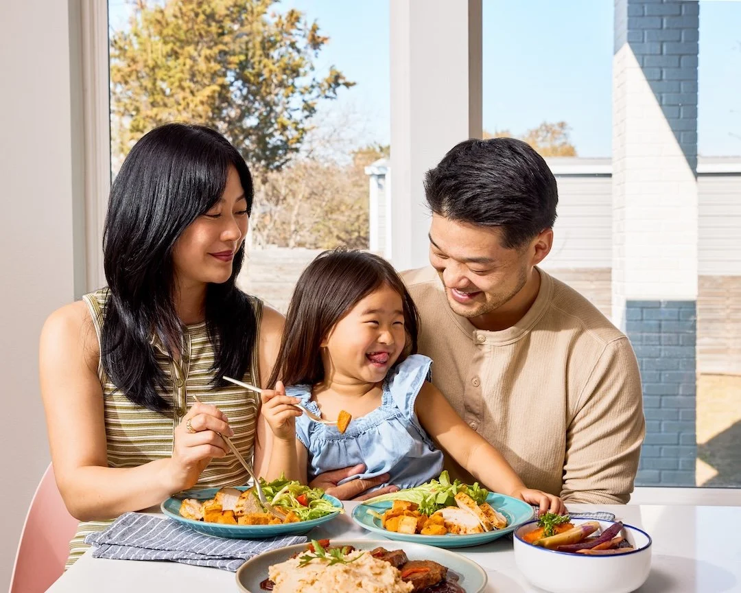 A family of three enjoying a meal together at a table near a window, with a woman, a man, and a young girl smiling and sharing food.