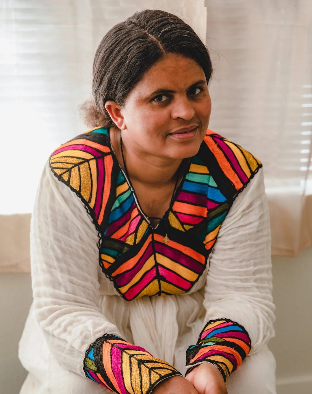 A woman with dark hair sitting indoors, wearing a white garment with colorful embroidered patterns on the shoulders, cuffs, and chest.