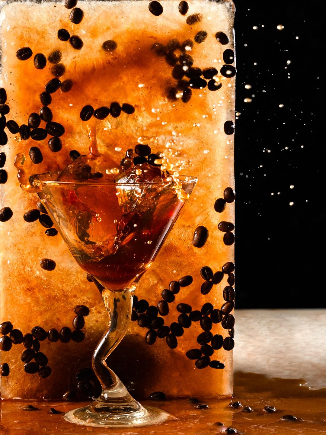 A glass of dark liquid with ice cubes falling into it, coffee beans scattered around, set against a textured beige and black background.