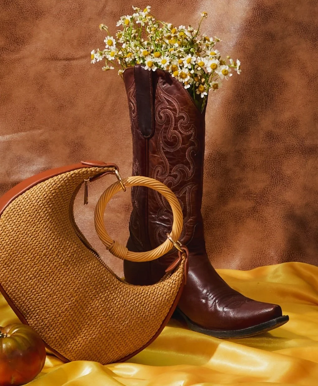 A tall brown leather cowboy boot filled with daisies, an orange woven handbag with a bamboo handle leaning against the boot, a tomato on a yellow satin fabric, and a textured brown background.