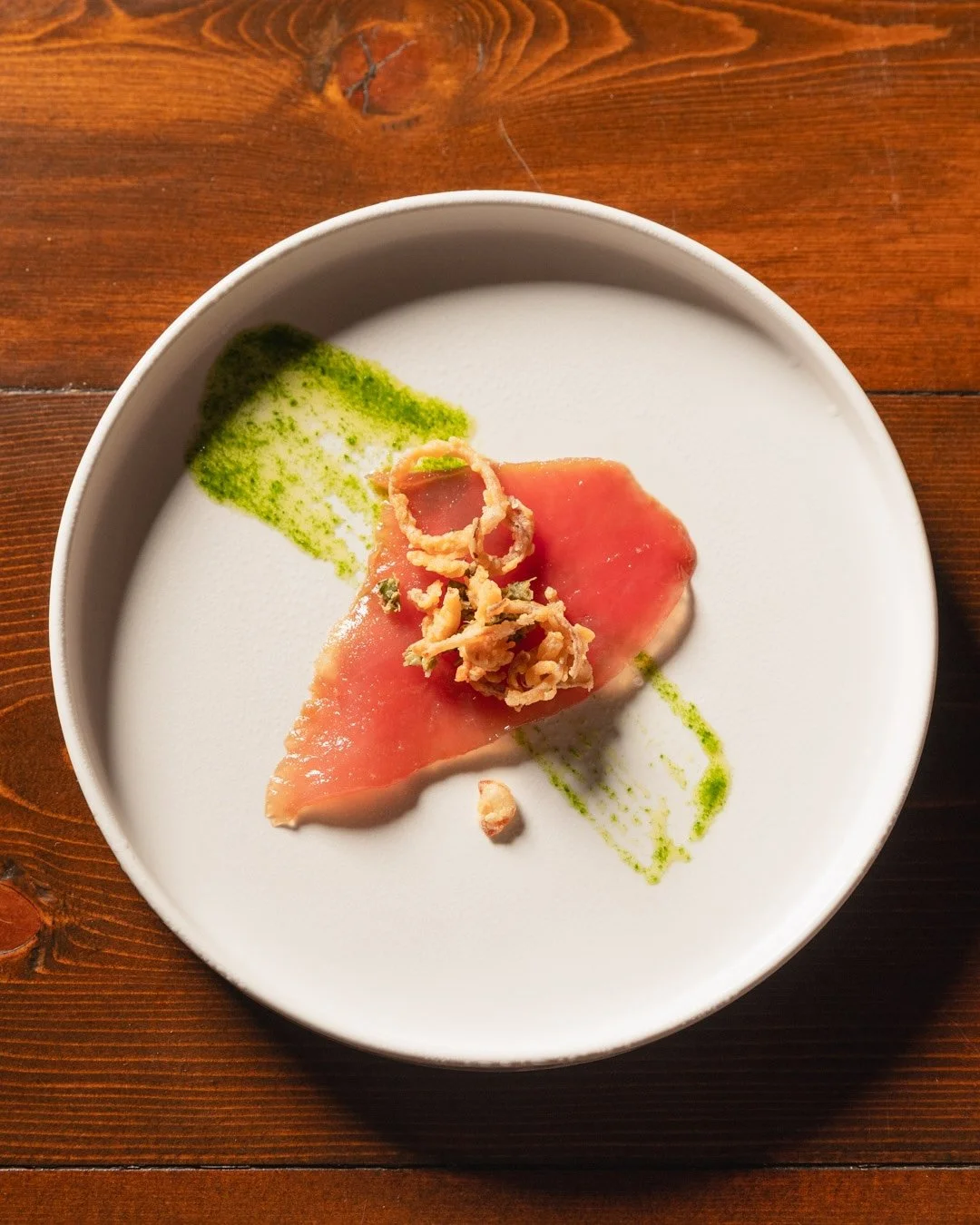 A plate with a slice of pink tuna sashimi garnished with crispy fried onion strips, a green sauce smear, and a small dollop of white cream on a wooden table.