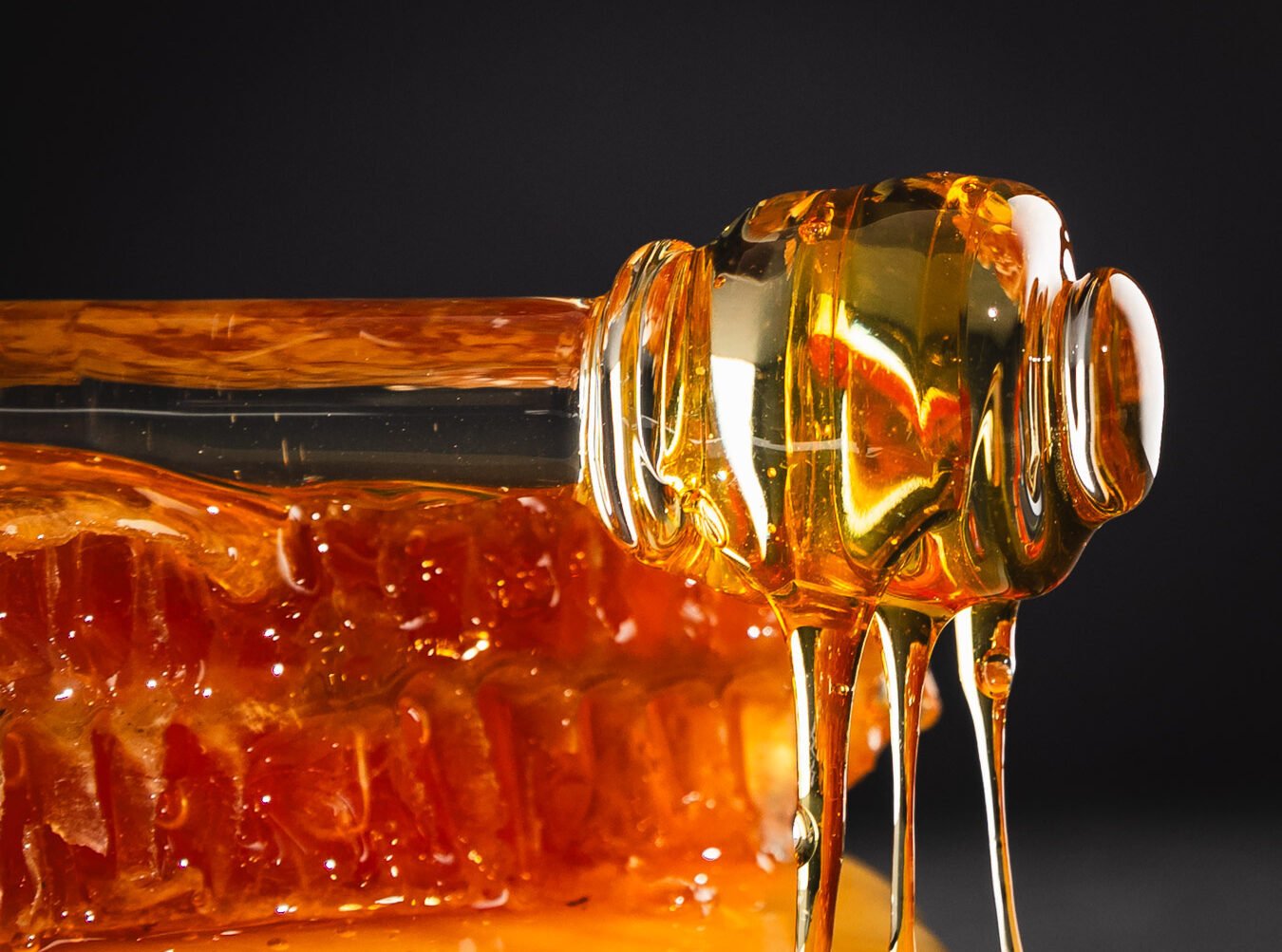 Close-up of honey dripping from a wooden honey dipper stuck in a honeycomb against a dark background.