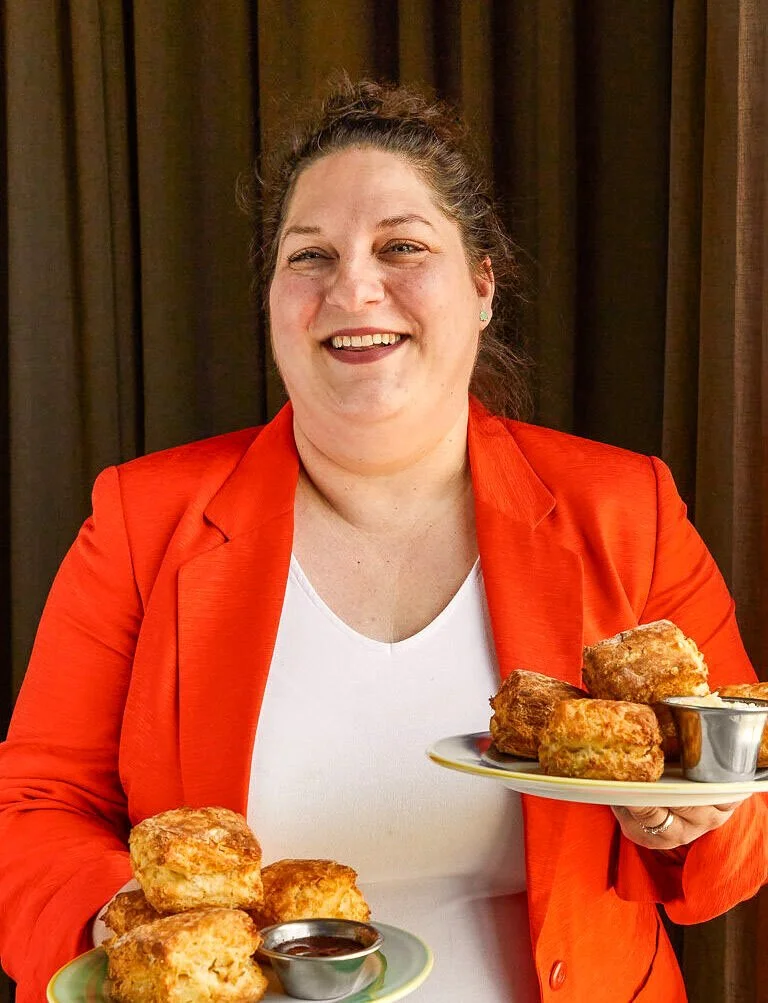 A smiling woman in an orange blazer holding two plates of biscuits and jam, standing in front of a dark brown curtain.