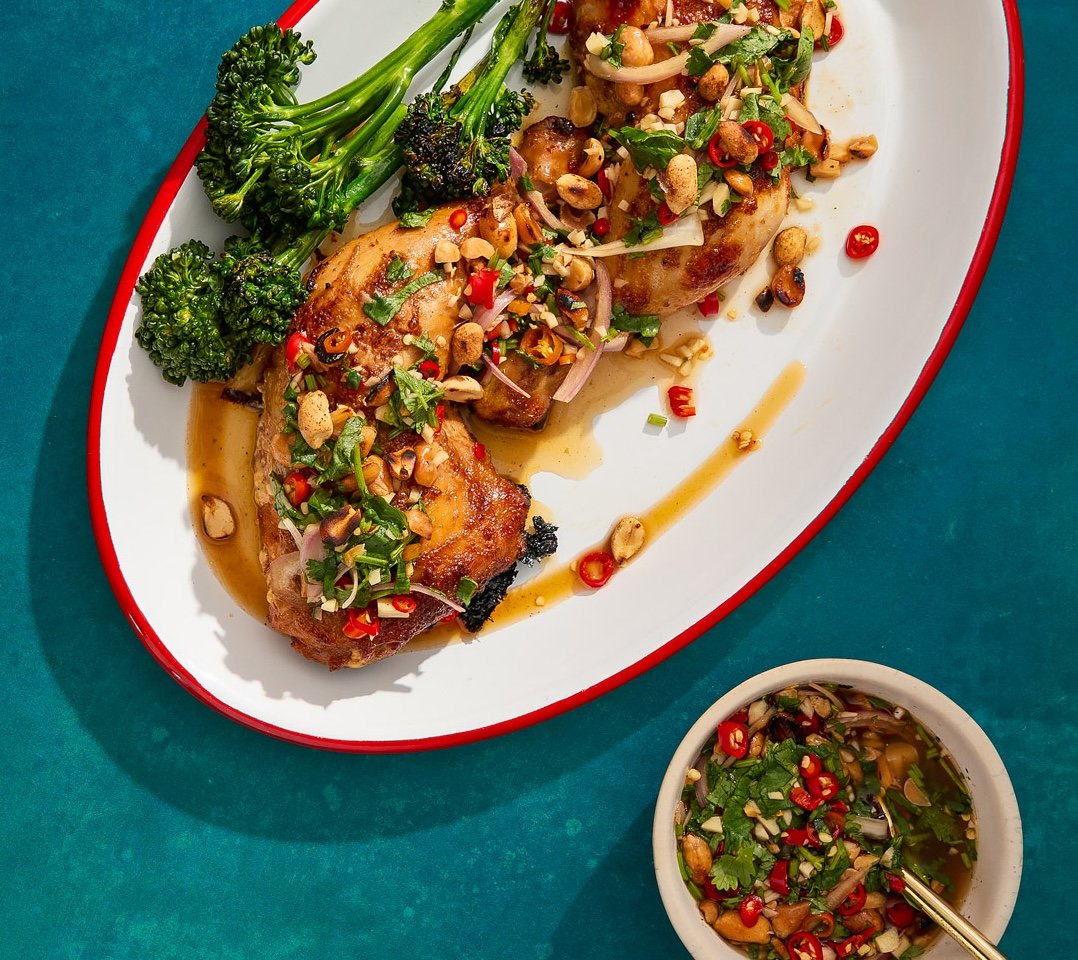 Grilled chicken thigh topped with chopped peanuts, red chili slices, cilantro, and green onions, served with broccolini, and a small bowl of dipping sauce with chopped herbs and red chili slices.