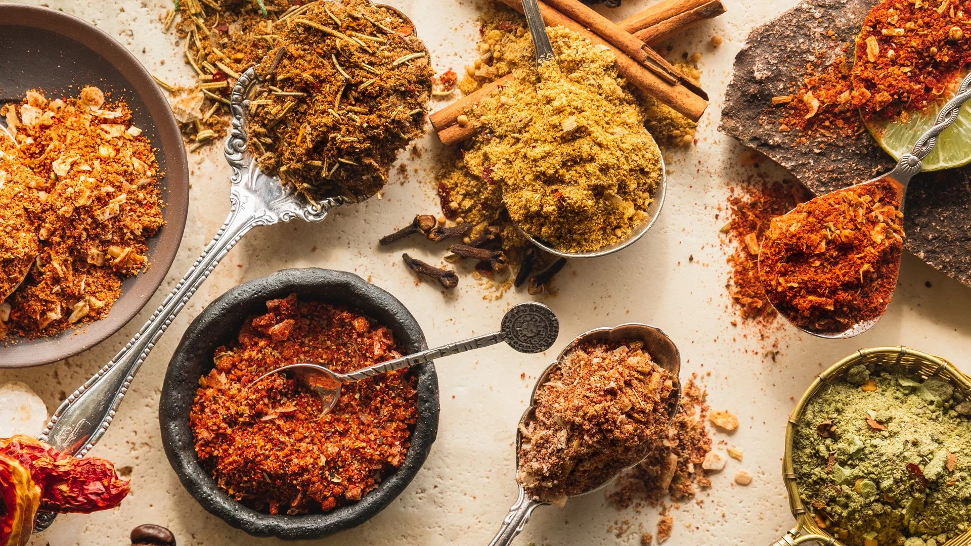 Assorted spices and herbs in bowls and on a surface, including dried chili flakes, ground turmeric, cinnamon sticks, and other seasonings.