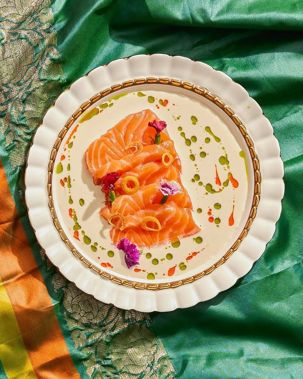 Sliced salmon sashimi garnished with edible flowers, thin slices of lime, and colorful sauce drizzles on a white plate with a gold rim, set on a colorful, patterned fabric background.
