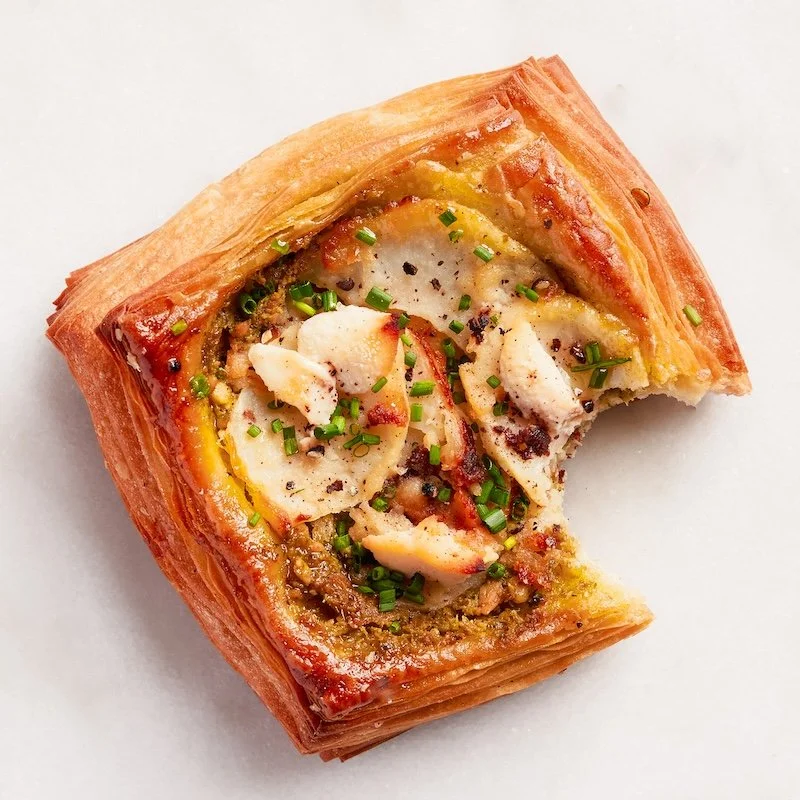 A square piece of savory pastry with a golden-brown flaky crust, filled with sliced bananas, nuts, and green herbs, topped with black pepper.
