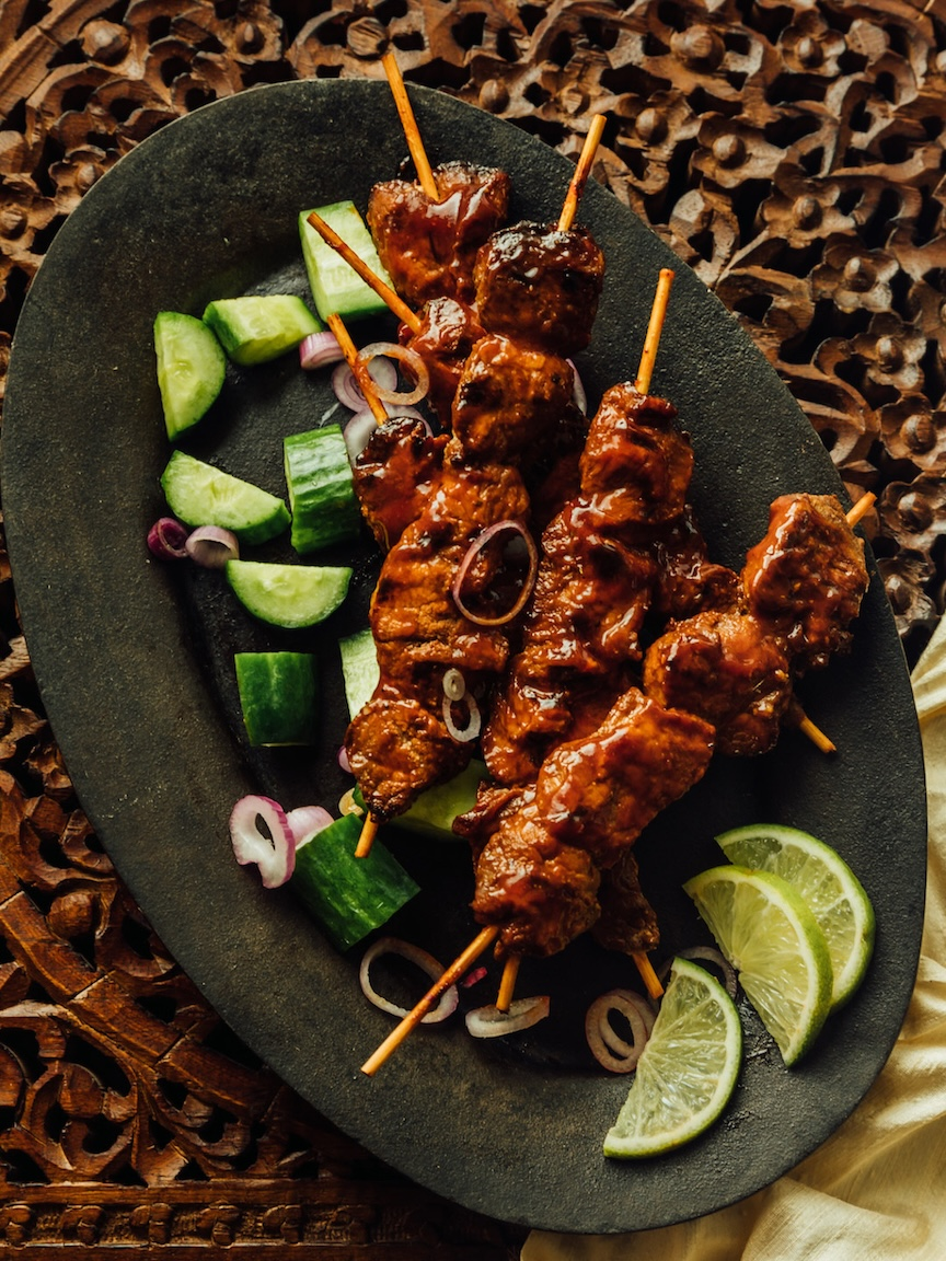 Skewers of grilled meat with barbecue sauce, garnished with cucumber chunks, onion slices, and lime wedges on a black plate.