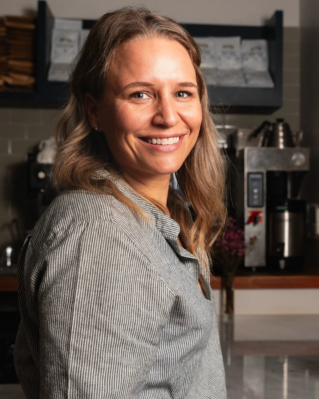 A smiling woman with wavy, shoulder-length hair in a kitchen setting, wearing a striped shirt.