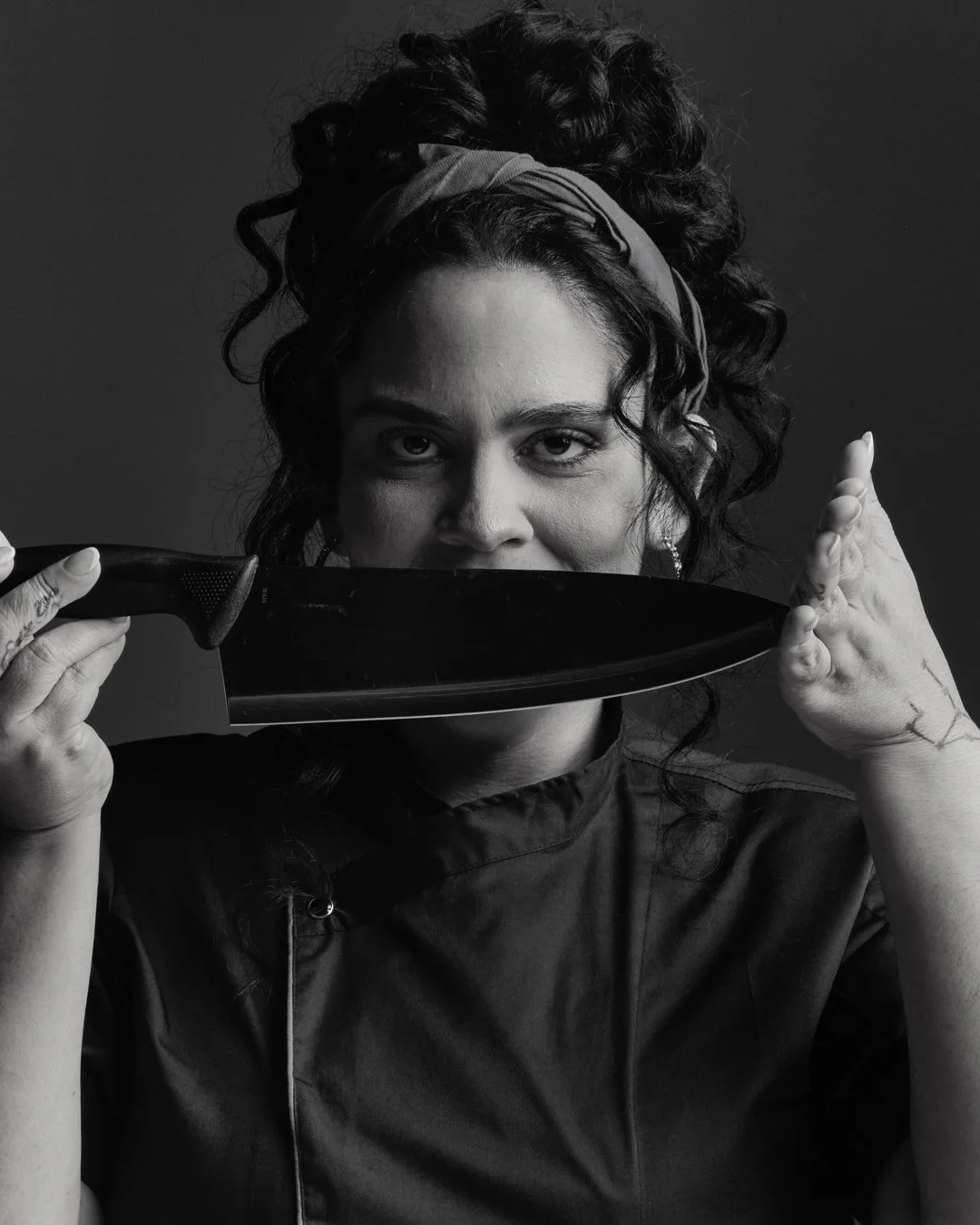 Black and white photo of a woman with curly hair, wearing a headband and earrings, holding a large kitchen knife partially covering her face, with her eyes staring directly at the camera.