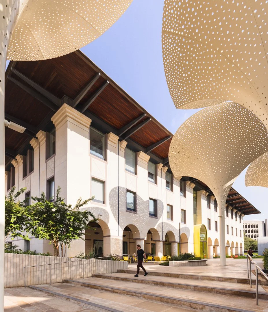 Modern building with arched openings and decorative perforated canopy structures overhead, casting patterned shadows on the facade and ground during daytime.