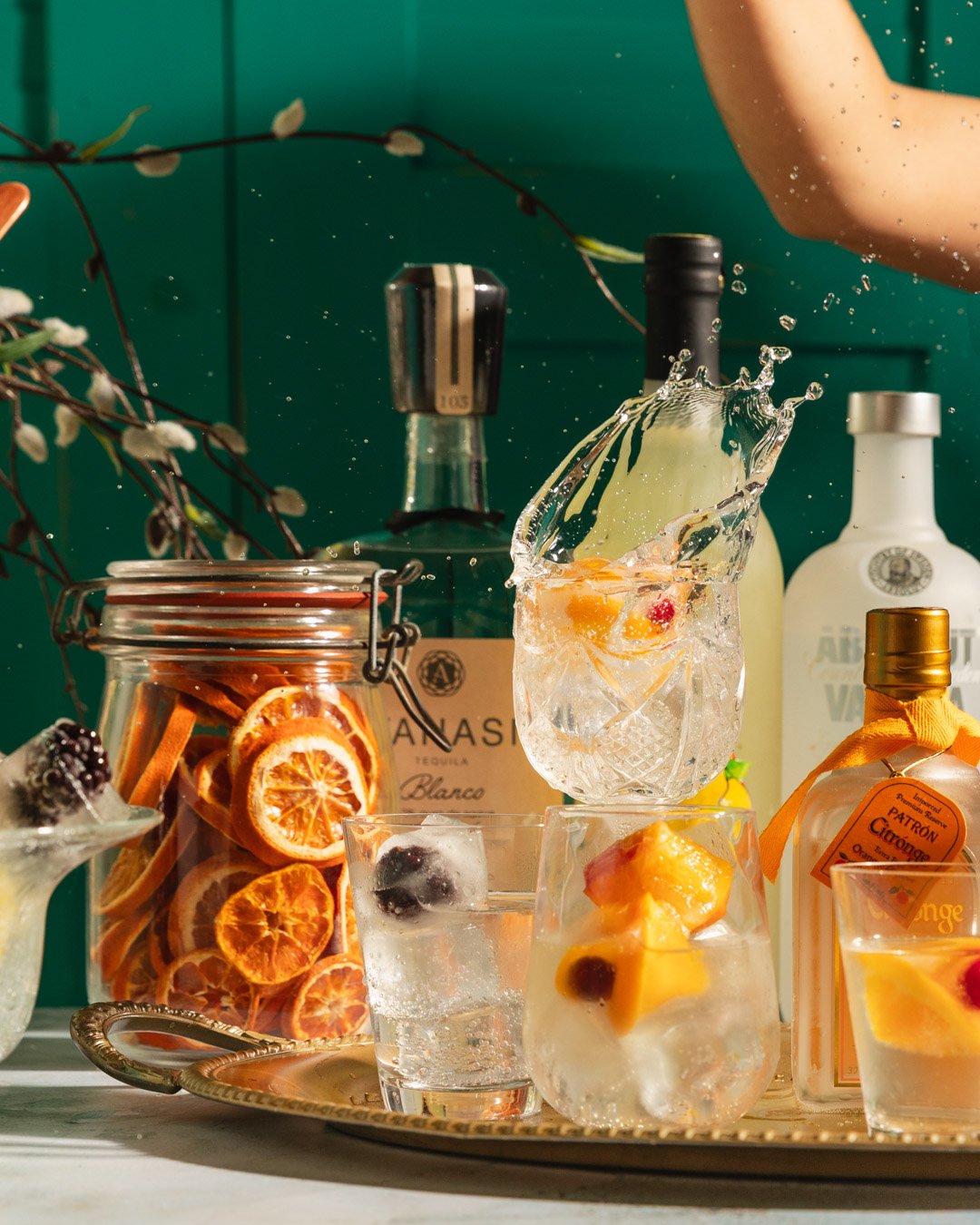 A collection of cocktails and infused water with citrus slices and berries on a decorative tray, with bottles of alcohol and garnishes in the background.
