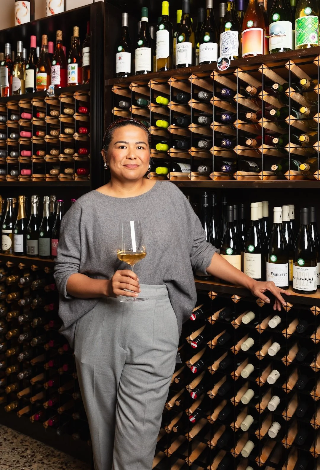 A woman standing in front of a wine rack, holding a glass of white wine and smiling.