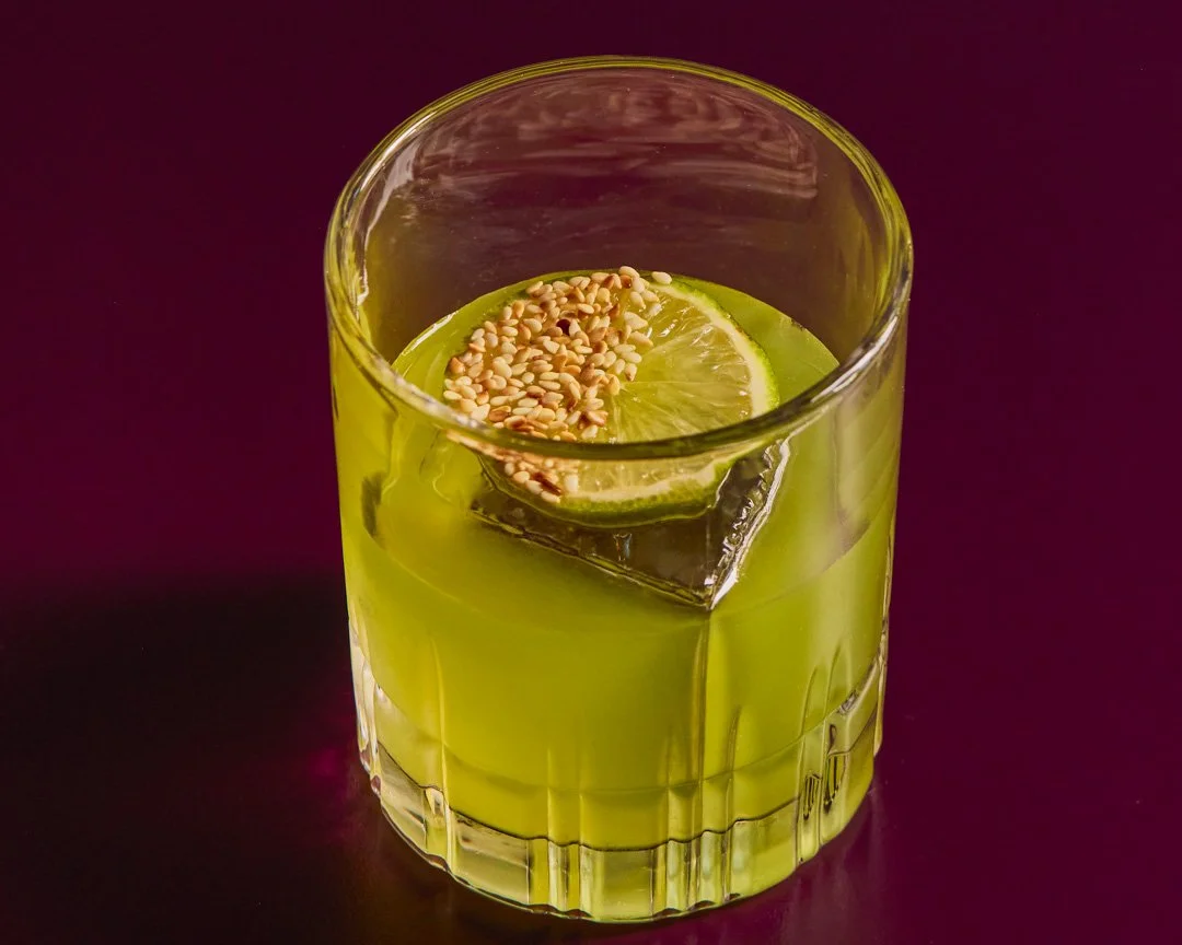 A glass of green juice with a lemon slice topped with sesame seeds on a dark purple background.