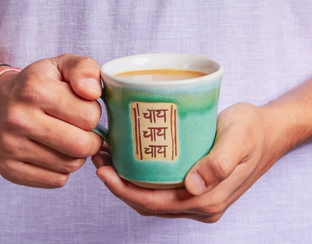 A person holding a light green ceramic mug filled with a hot beverage, with Hindi script written on the mug, against a purple background.