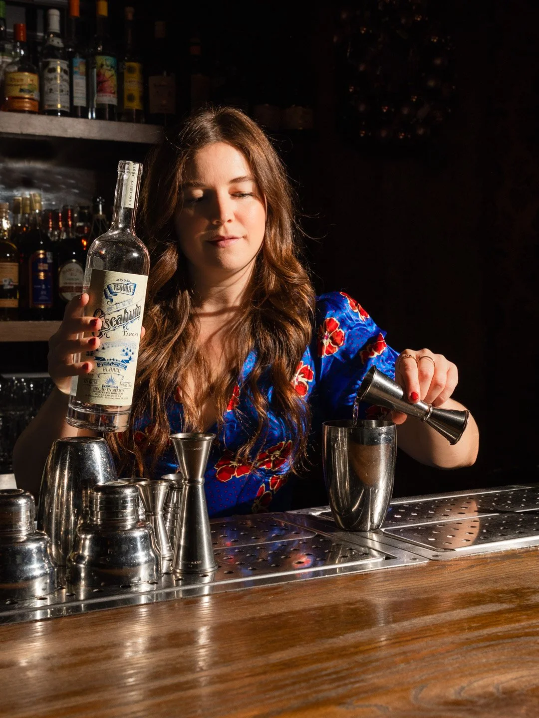 A woman behind a bar pouring a liquid into a shaker, holding a bottle of tequila, with bar tools and bottles on shelves in the background.