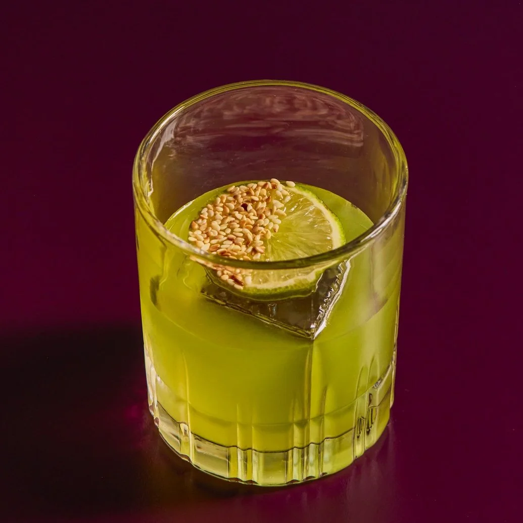 A glass of green juice with a slice of lime and sesame seeds on top, placed on a dark surface.