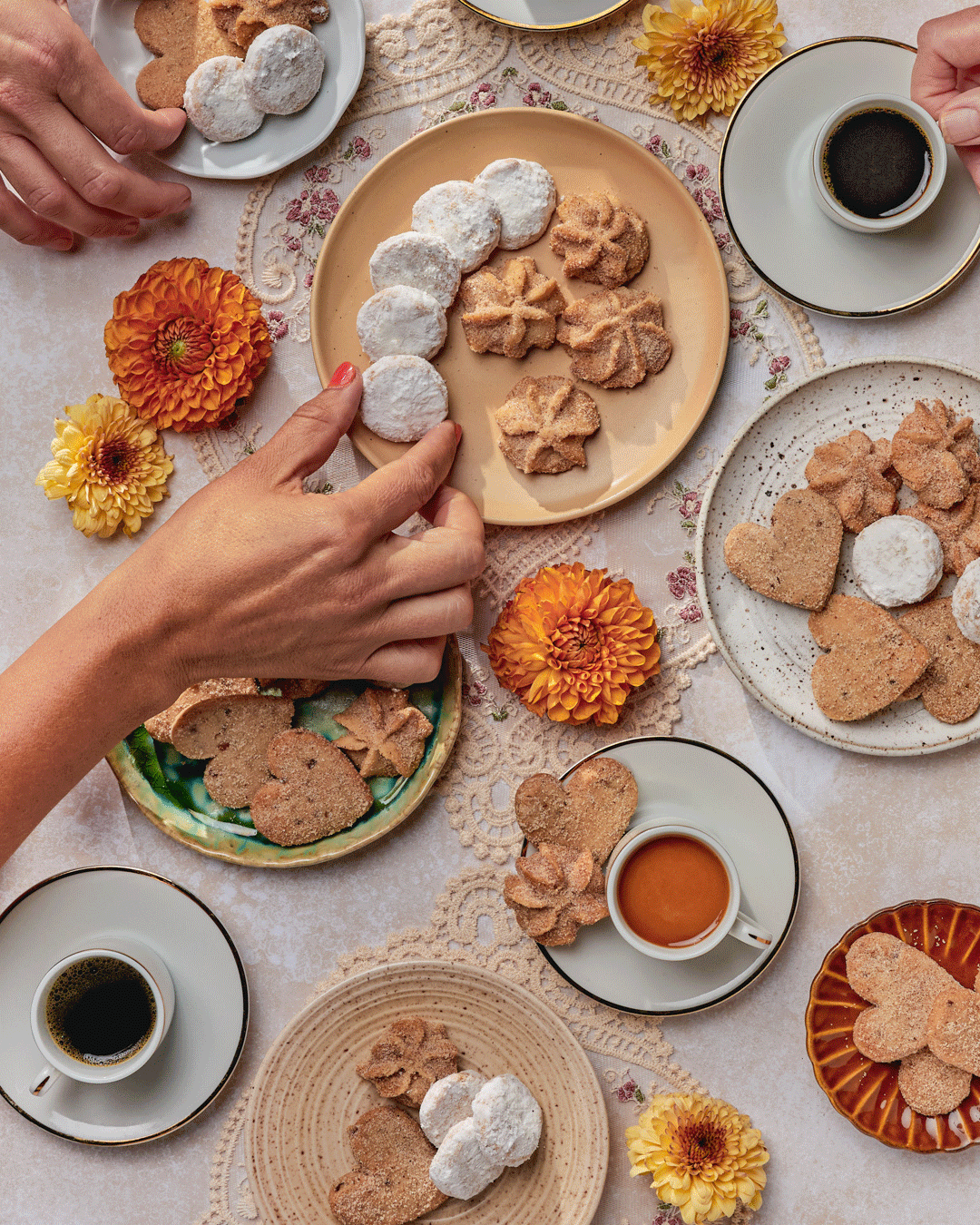 An overhead view of a table with various cookies, coffee cups, and flowers. A person is reaching for cookies on a plate.