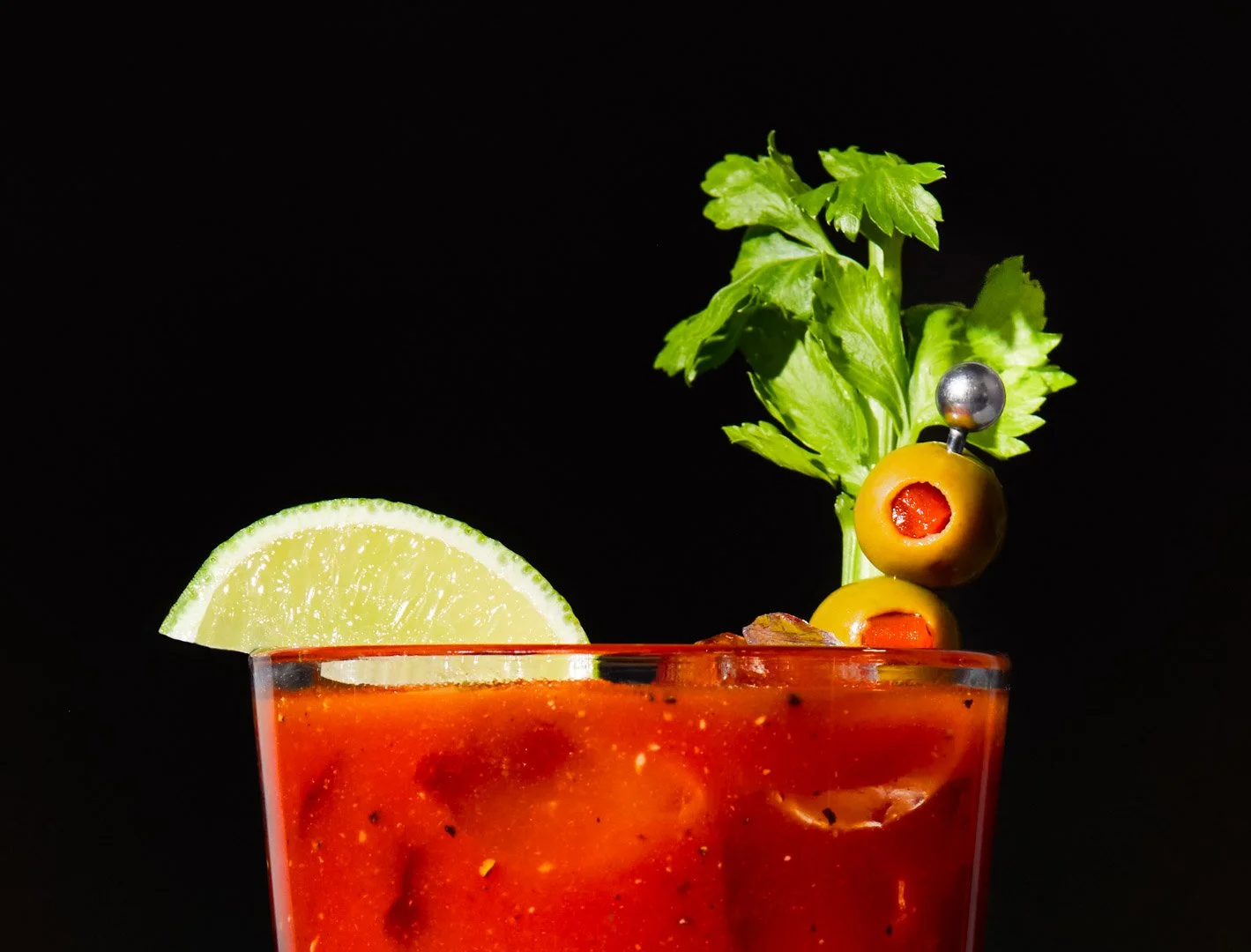 Close-up of a Bloody Mary cocktail in a glass, garnished with a lime wedge, green olives on a toothpick, a sprig of celery, and a black olive on a toothpick, against a black background.