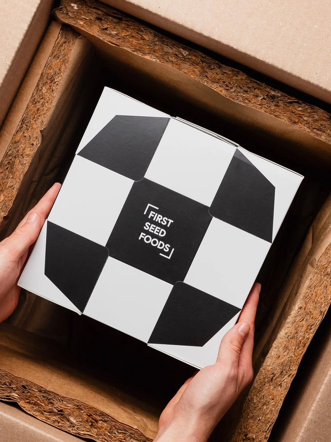 A person holding a black and white box labeled 'First Seed Foods' inside a cardboard shipping box.
