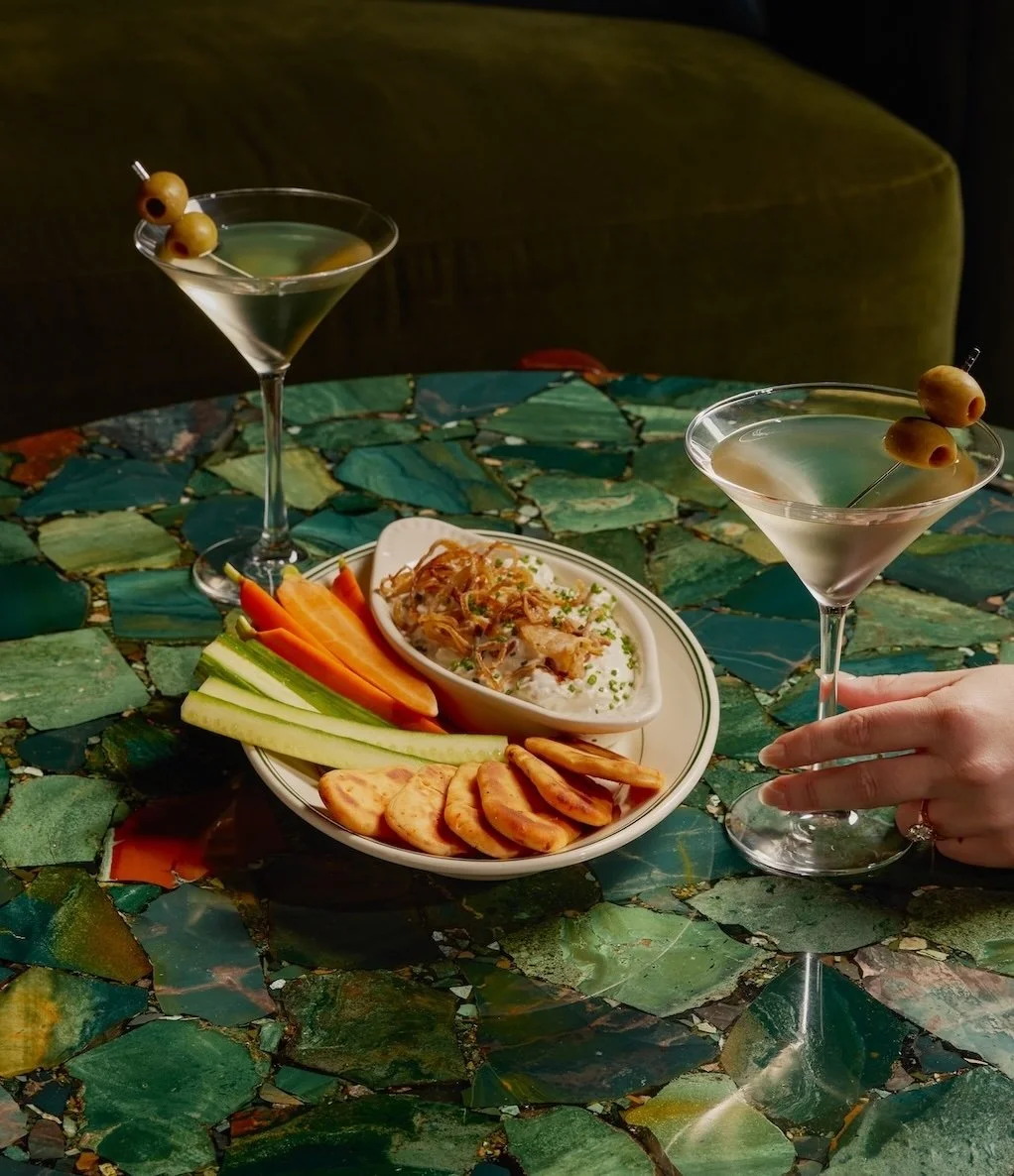 Two martini glasses with olives, a plate of sliced vegetables, a bowl of a dish with shredded meat and rice, and a hand holding one of the martini glasses on a green mosaic table.