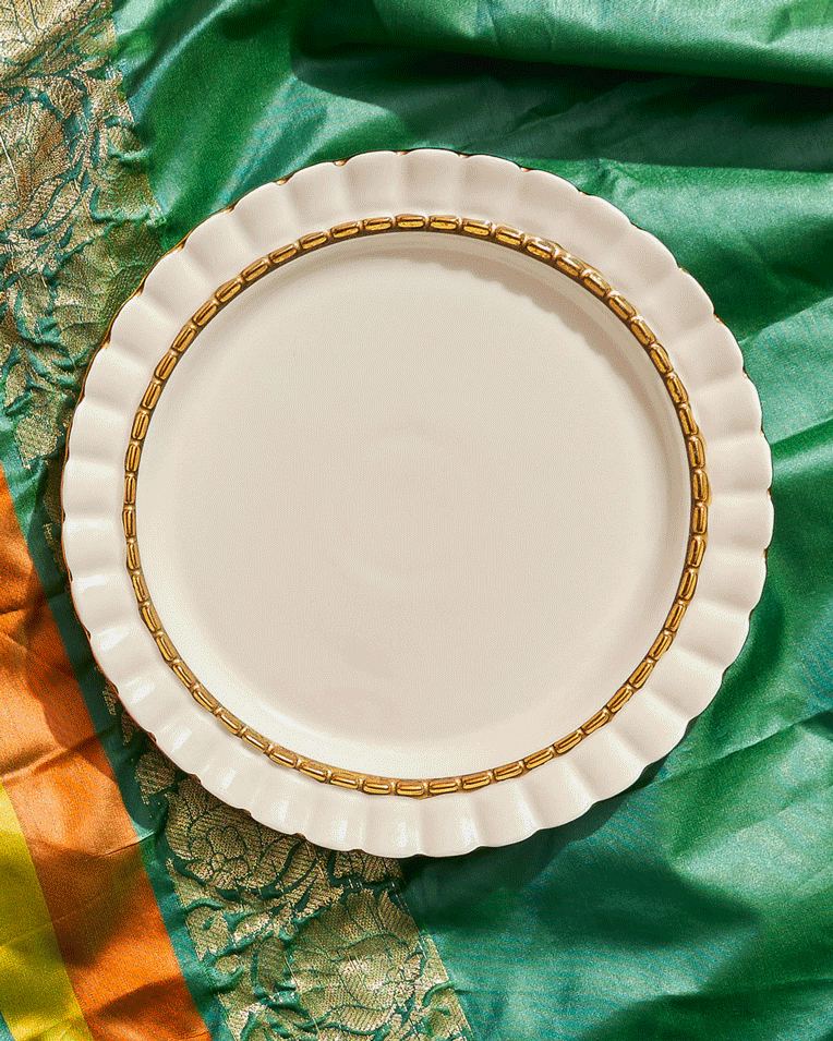 A decorative white plate with a gold-rimmed scalloped edge, placed on colorful fabric with green, orange, and gold details.
