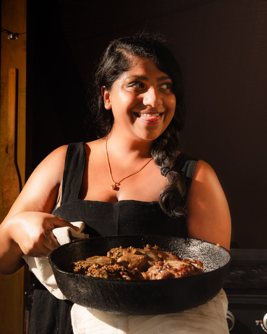 A woman with dark hair in a braid, wearing a black sleeveless top and gold necklace, smiling while holding a black cast iron skillet with cooked food inside.