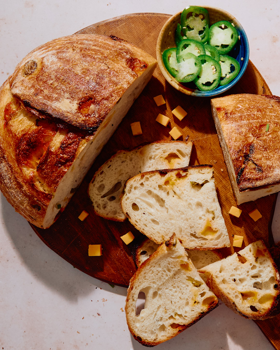 Loaf of sliced Italian bread with cheese, a small bowl of sliced jalapenos, and small cheese cubes on a wooden platter.