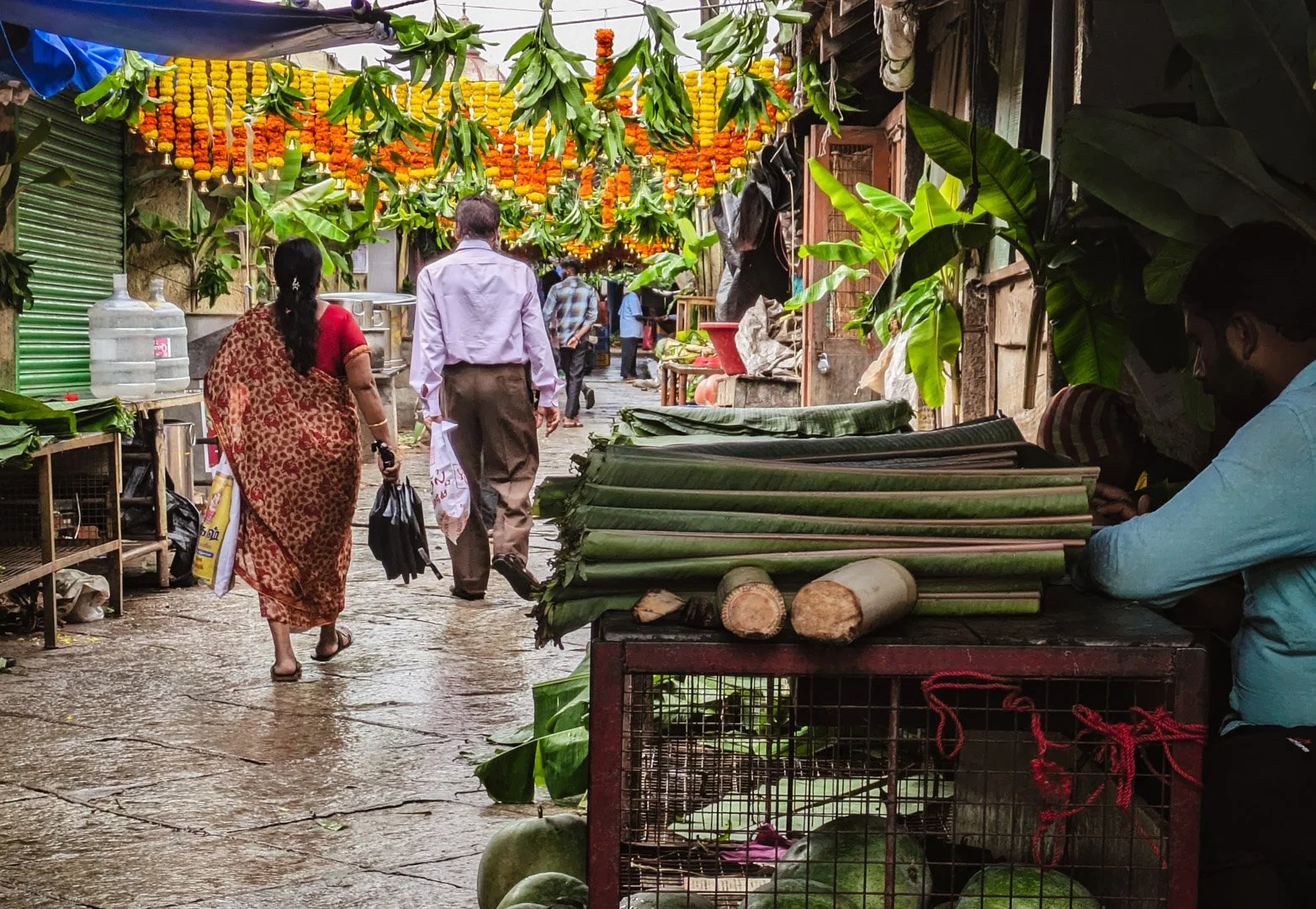 A busy street market with people walking, vendor stalls with green banana leaves, and hanging marigold flower garlands overhead. One vendor is sitting behind a cart with long green vegetables, while other shoppers walk through, some carrying umbrella