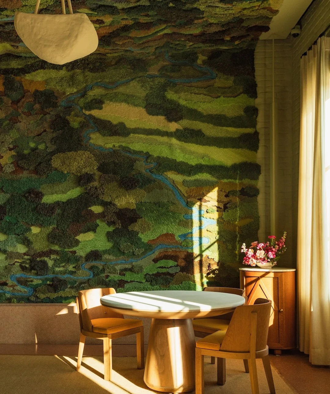 A round table with chairs in a room illuminated by natural sunlight, which casts shadows across the table. A colorful, textured mural on the wall behind depicts a landscape with rivers and forests. To the right, a cabinet with a bouquet of pink flowers on top, near a large window with cream-colored curtains.