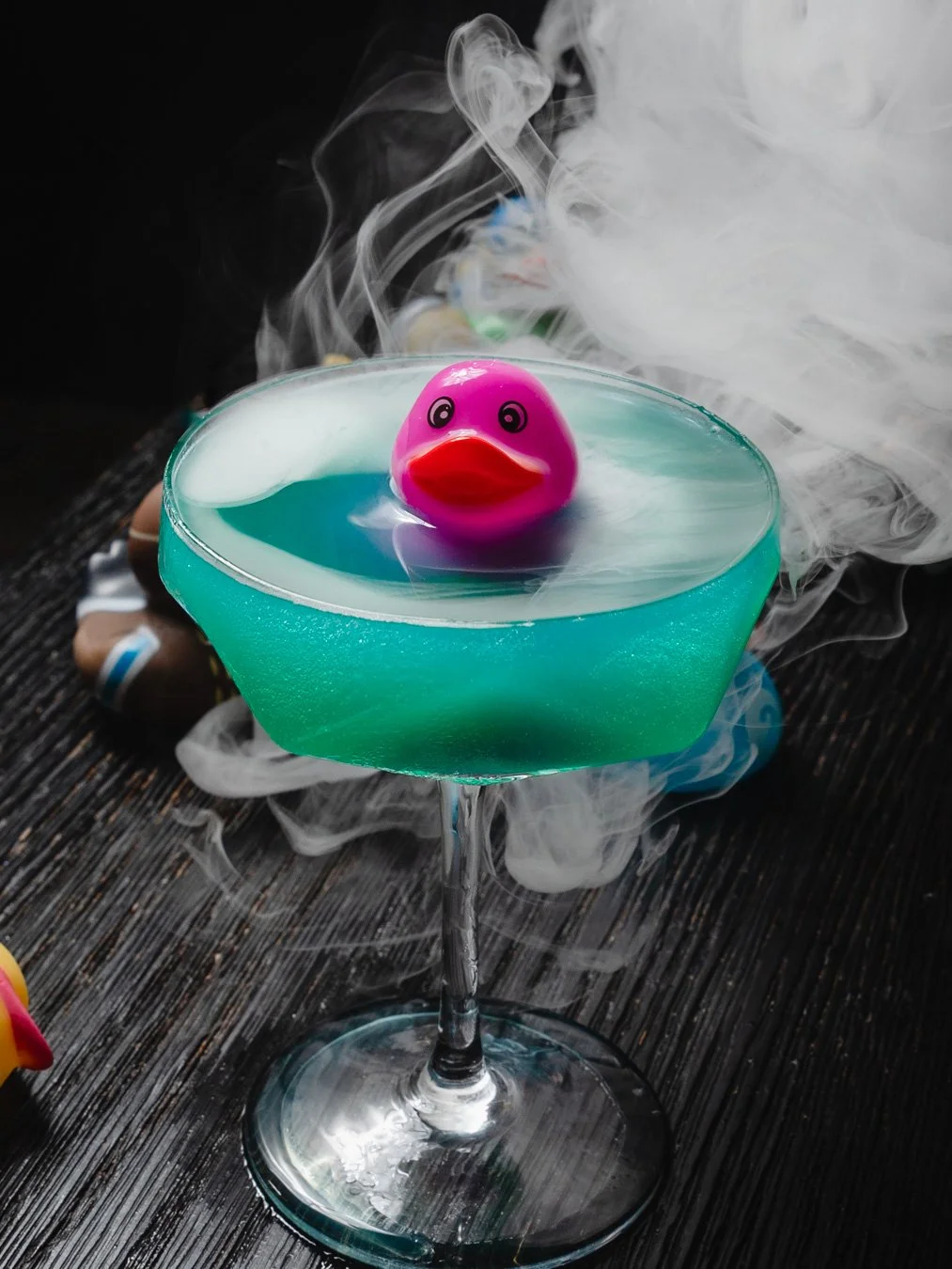 A cocktail glass with a blue drink, topped with a rubber duck with exaggerated lips, surrounded by smoke and a dark wooden surface.