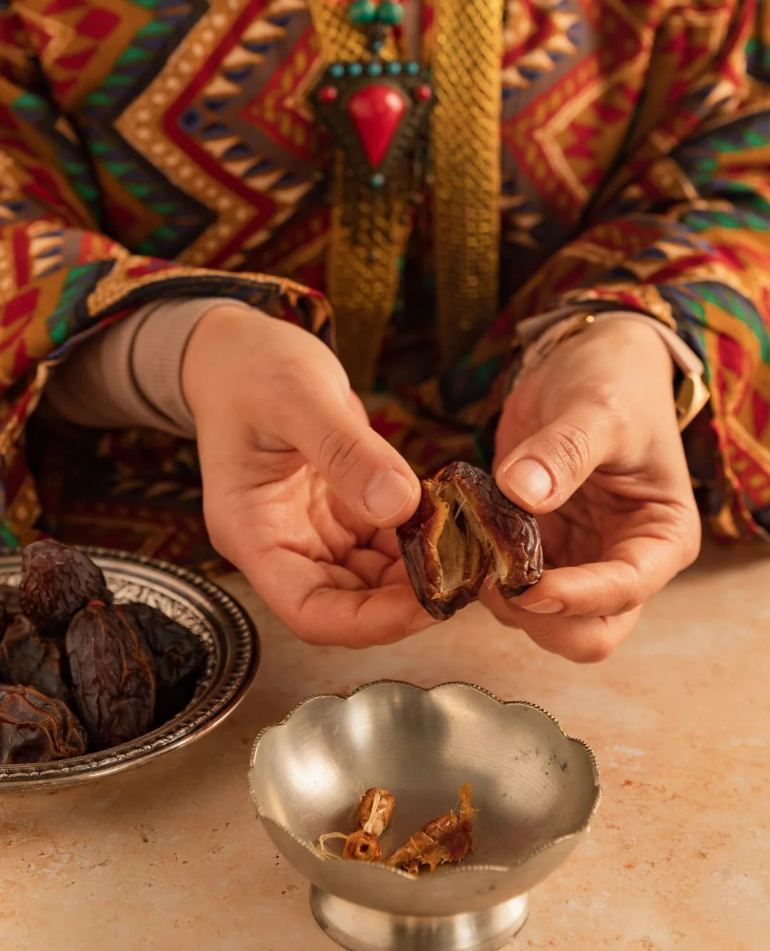 Person wearing a colorful, patterned jacket holding a opened date fruit, with a bowl of dates and date parts on a table.