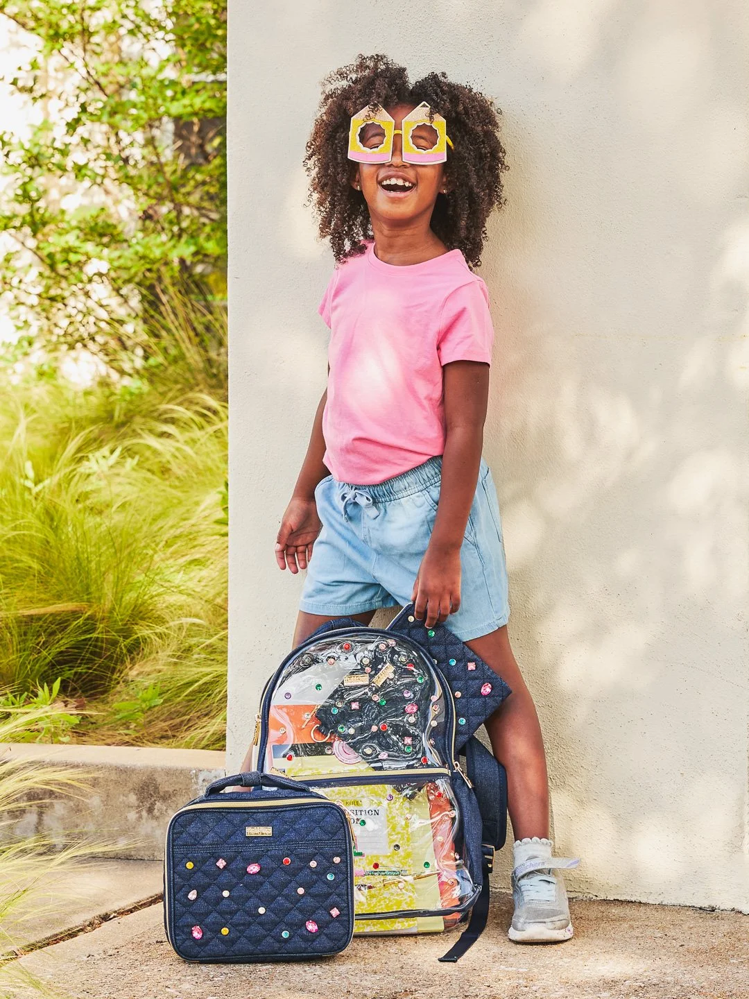 A smiling young girl with curly hair wearing pink sunglasses, a pink t-shirt, and light blue shorts, standing outdoors against a light-colored wall with greenery in the background, holding a decorated backpack.