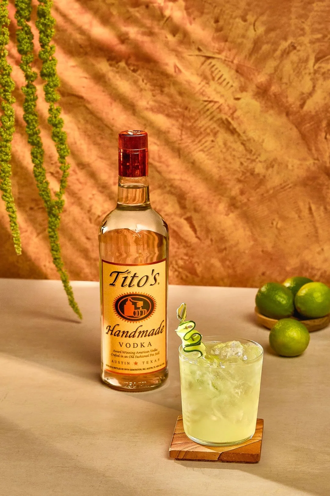 A bottle of Tito's Handmade Vodka and a glass of a lime-colored cocktail with ice, garnished with a curled lime peel, on a light-colored table with a wall, some hanging greenery, and a small dish of limes in the background.