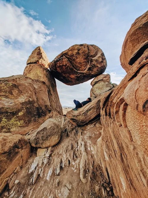 Person relaxing among large, weathered rocks and boulders outdoors with a partly cloudy sky.