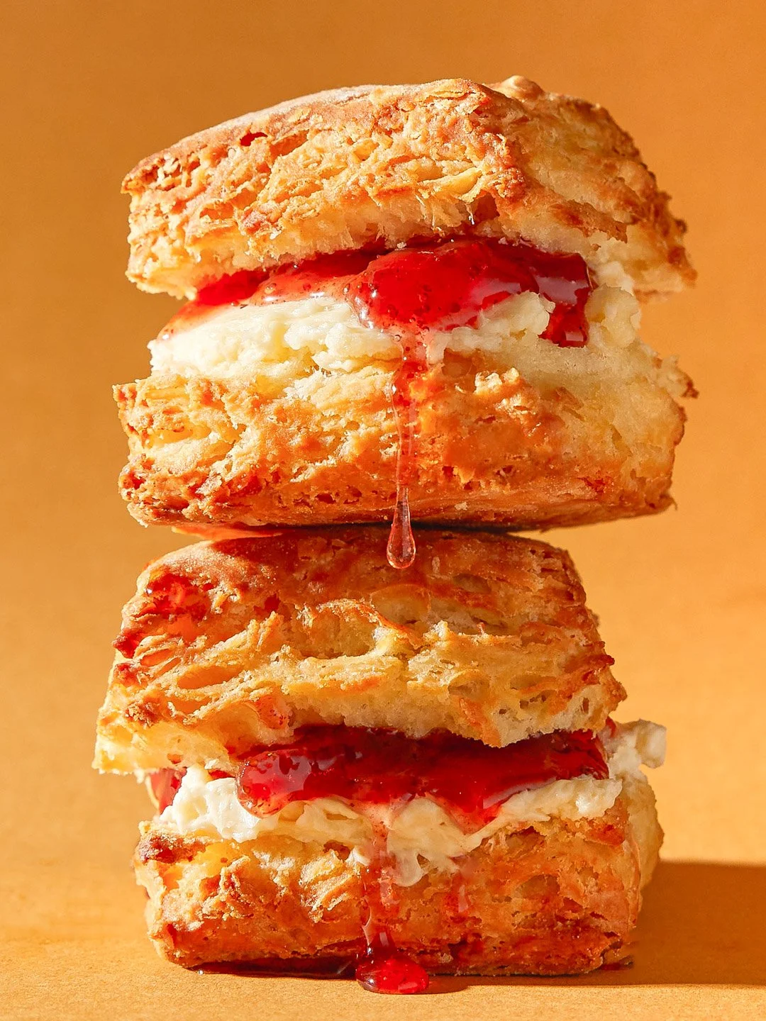 Stacked scones with cream and strawberry jam, with some jam dripping down, on a wooden surface.