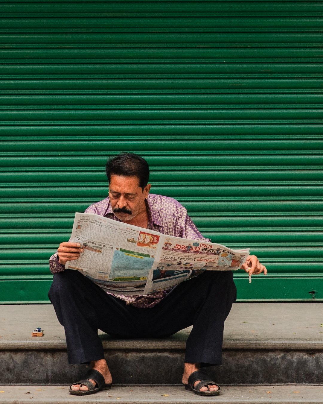 Man sitting on sidewalk reading a newspaper in front of a closed green shutter door.