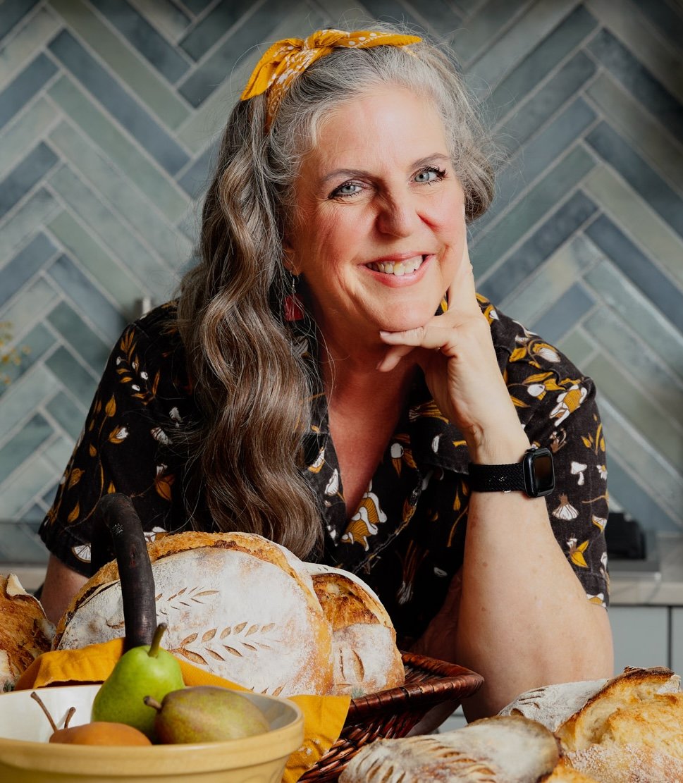 A smiling woman with gray hair, wearing a yellow headband and a black shirt with a leaf pattern, sitting at a table filled with bread and fruit, in a kitchen with a gray tile backsplash.