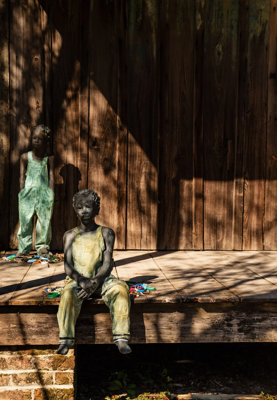 Two life-sized sculptures of children, one sitting and one standing, on a wooden porch in front of a wooden fence, with scattered toys and shadows from nearby trees.
