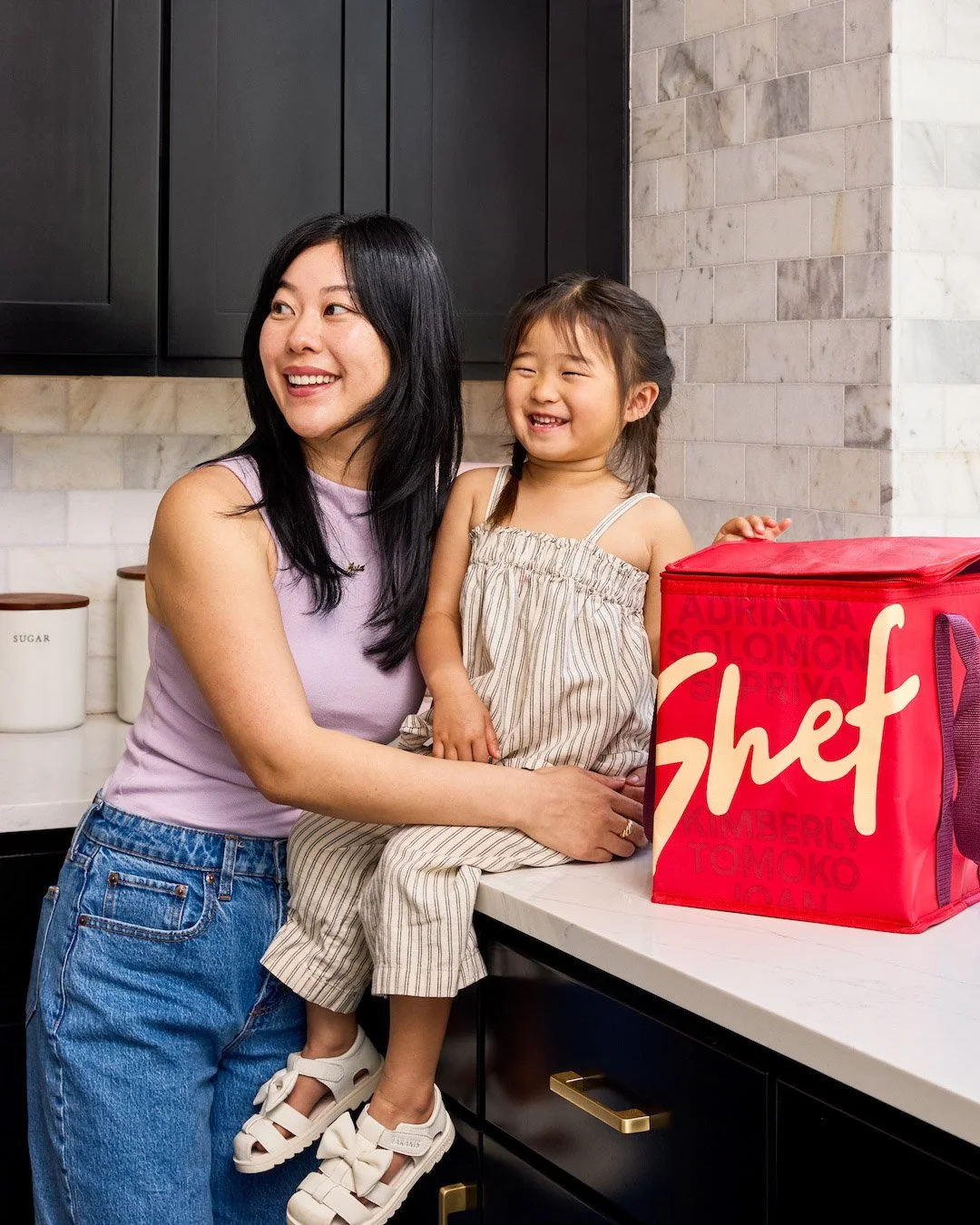 A woman and her young toddler are smiling in a modern kitchen, with a red insulated bag on the white countertop. Product photography by San Francisco Bay Area photographer Nitya Jain.